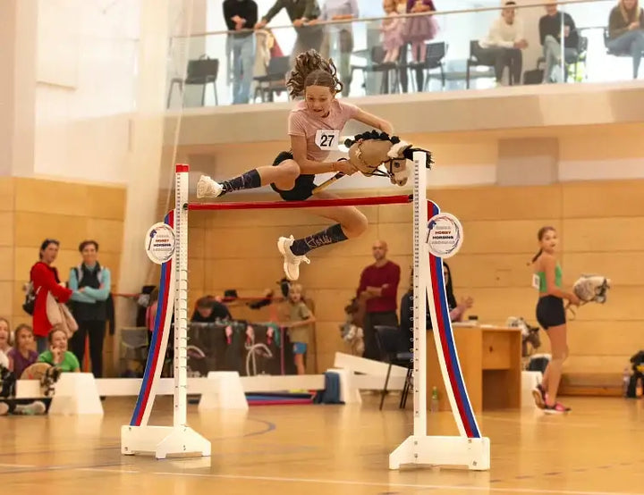 Tekmovalec v športu Hobby Horsing s Hobby Horse konjem preskakuje rdeč drog na dogodku v dvorani