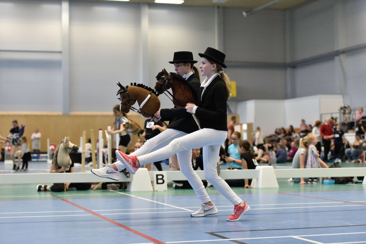Dva otroka v svečanih tekmovalnih oblačilih za Hobby Horsing tekmujeta s Hobby Horse konji v dvorani