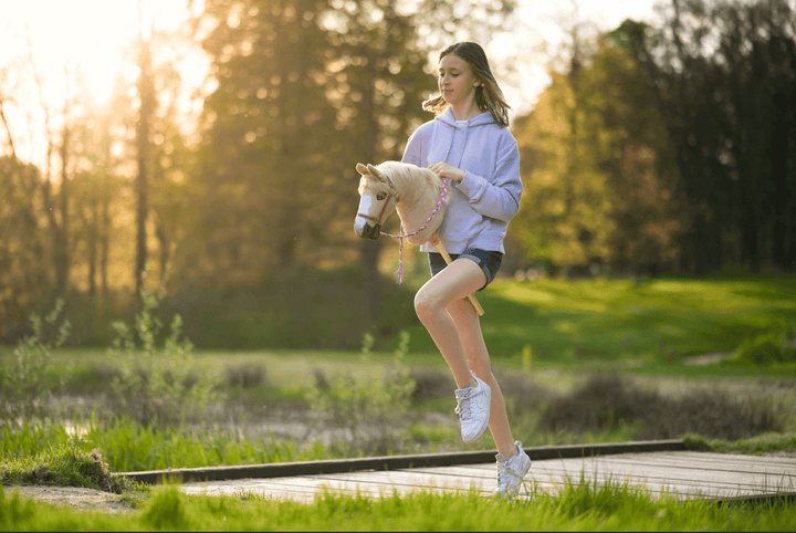 Deklica ob sončnem zahodu na prostem uživa v športu Hobby Horsing z belim Hobby Horse konjem