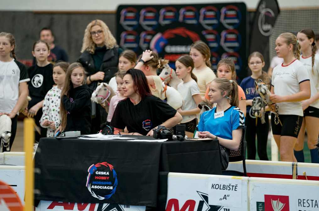 Živahen prizor z dogodka Hobby Horsing v dvorani: dva organizatorja sedita za črno mizo z logotipom Češke zveze Hobby Horsing (ČHHA), okoli njiju stojijo dekleta v športnih oblačilih s Hobby Horse konji. Vidne so bele ograje arene in napis Kolbiště »A«.