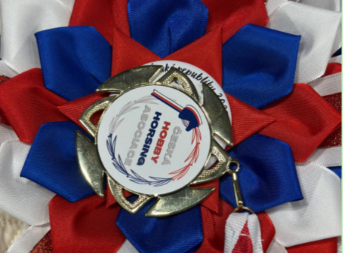 Bližnji posnetek rdeče-belo-modre nagradne rozete z zlato medaljo in emblemom Hobby Horsing v sredini, praznična podrobnost s češkega prvenstva Hobby Horsing 2025.