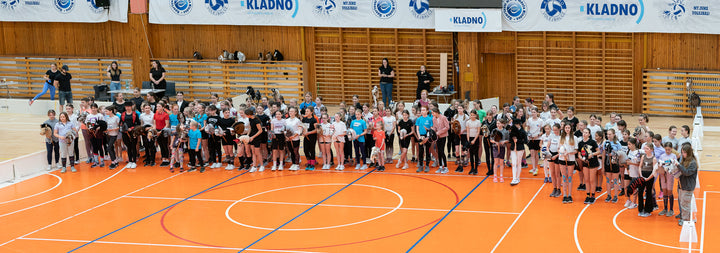 Velika skupina mladih tekmovalcev v športu Hobby Horsing v športnih oblačilih stoji v vrsti na oranžnem igrišču v dvorani in drži različne Hobby Horse konje, v ozadju so leseni letveniki, tribune in transparenti Kladna.
