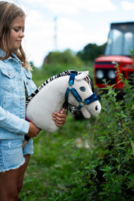 Deklica v jeans jakni drži Hobby Horse konja Aurora v modri oglavki na prostem ob zelenih rastlinah, zadaj rdeč traktor