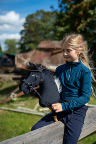 Svetlolasa deklica v modrozeleni majici sedi na leseni ograji in drži Hobby Horse konja Bond s črno oglavko, ki ima tukaj črno uzdo z vajetmi