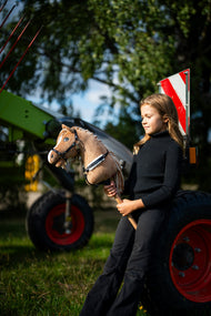 Deklica v črnem puloverju z visokim ovratnikom ob zelenih kmetijskih strojih drži Hobby Horse konja Bren s črno oglavko, tu s črno uzdo