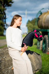 Deklica v smetanastem puloverju sedi na veliki skali na prostem s črnim Hobby Horse konjem v črni oglavki z roza krznom