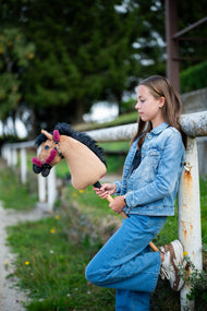 Deklica v jakni iz džinsa, naslonjena na belo ograjo, drži svetlo rjavega Hobby Horse konja z malinasto oglavko s krznom