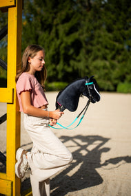Deklica v roza majici, naslonjena na rumeno stojalo ovire na jahališču, s Hobby Horse konjem Freya s črno-turkizno uzdo