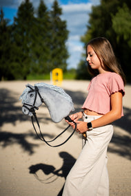 Deklica v roza majici hodi čez peščeno jahališče s Hobby Horse konjem Grace s črno uzdo in vajetmi