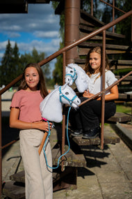 Dve deklici na zunanjem kovinskem stopnišču držita bela Hobby Horse konja s svetlo modrima oglavkama, eden ima povodec