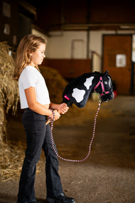 Deklica v beli majici stoji v hlevu s Hobby Horse konjem Merlin s črno oglavko, ki ima tukaj roza oglavko in pleten povodec