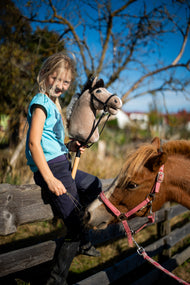 Nasmejana svetlolasa deklica na leseni ograji drži bež Hobby Horse konja v črni uzdi poleg ponija lisjaka v roza oglavki