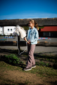 Mlada deklica v jakni iz džinsa ob ograji iz hlodov drži Hobby Horse konja Pixie s sivo oglavko s krznom in povodcem