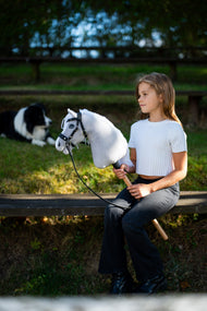 Deklica v beli majici sedi na klopi s svetlo sivim Hobby Horse konjem s črno uzdo, za njo border collie