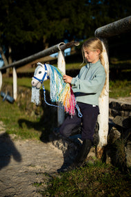 A girl in a mint sweatshirt by a white arena rail holds a white Hobby Horse with a rainbow mane on the blue striped lead rope