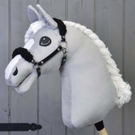 Siv plišast Hobby Horse konj s spleteno belo grivo nosi črno oglavko s črnim krznom, pred sivo leseno steno