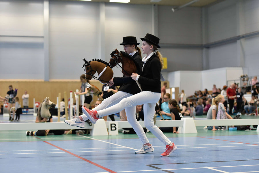 Dva otroka v svečanih tekmovalnih oblačilih za Hobby Horsing tekmujeta s Hobby Horse konji v dvorani