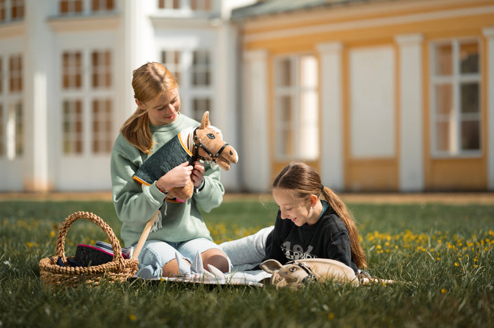 Deklica v svetlo zelenem puloverju sedi na travi ob pleteni košari z opremo za Hobby Horse konje in nežno drži svetlo rjavega Hobby Horse konja s črno uzdo, belo grivo in prešitim pregrinjalom. Ob njej je na odeji delno viden še en otrok.