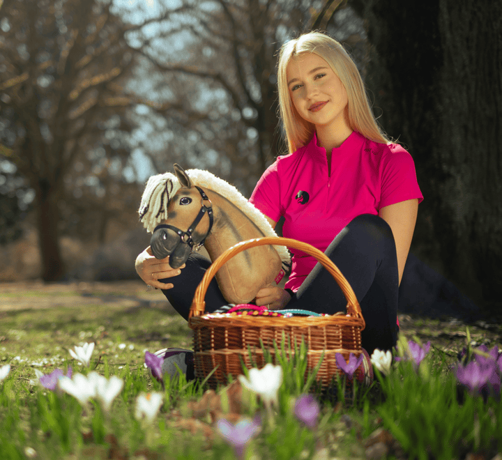 Mlada ženska v živo roza majici sedi zunaj na travi, v rokah drži Hobby Horse konja in pleteno košaro, polno pisanih jajc, obkrožena s cvetočimi belimi in vijoličnimi cvetlicami.