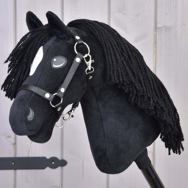 Črn plišasti Hobby Horse konj z dolgo grivo iz preje, belimi oznakami na glavi in črno uzdo, pritrjen na palico, pred svetlim lesenim ozadjem.
