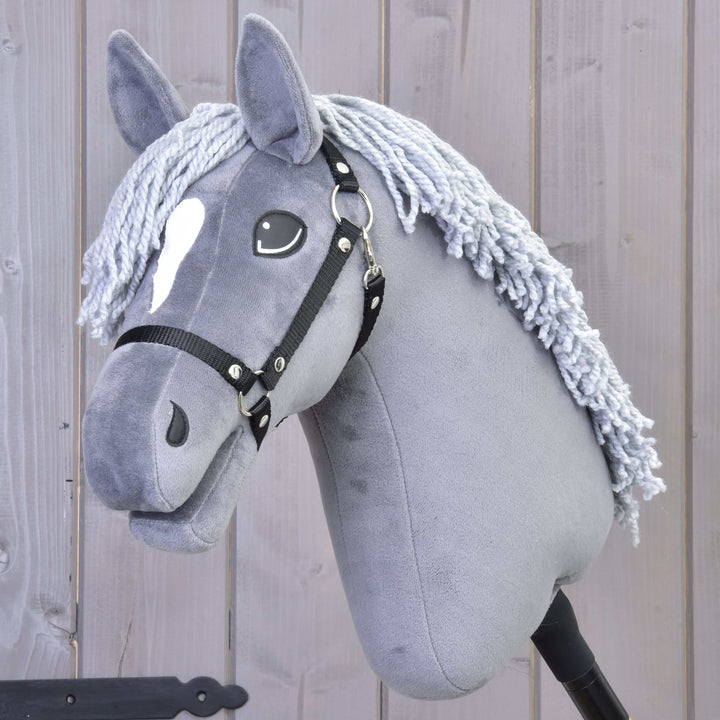 Siv Hobby Horse konj z belo pego na čelu, črno uzdo in grivo iz preje pred lesenim ozadjem.