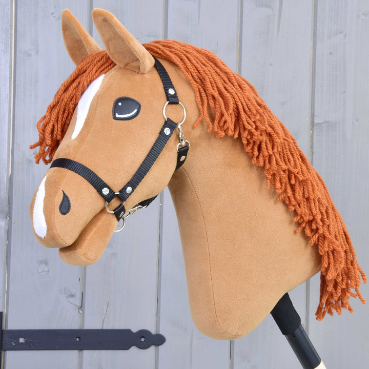 Ročno izdelan Hobby Horse konj z glavo iz rjavega blaga, belimi oznakami, črno uzdo in oranžno grivo iz preje, pritrjen na palico, pred sivim lesenim ozadjem.