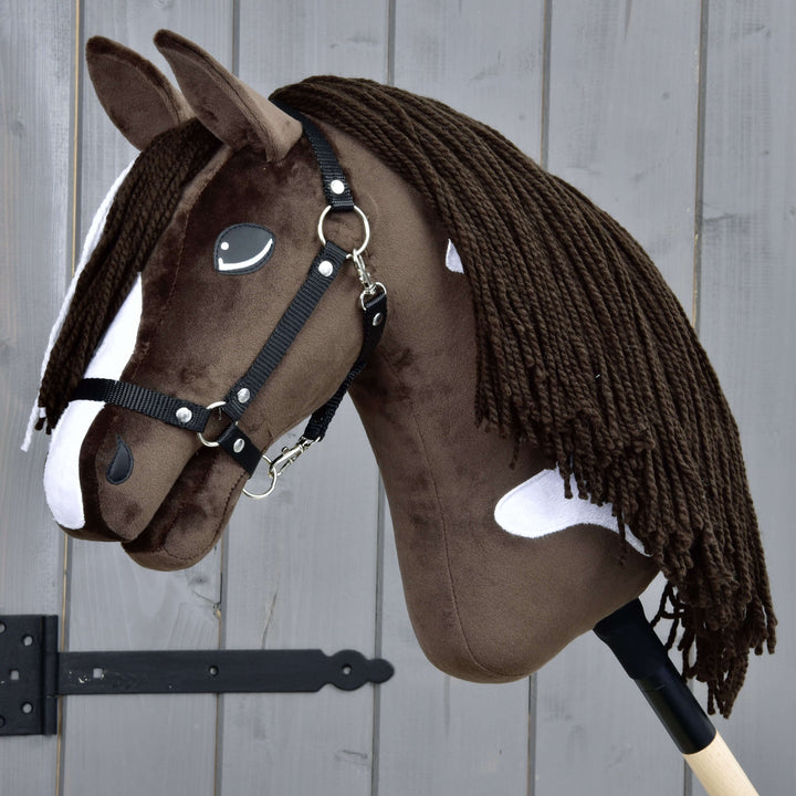 Realističen rjavo-bel Hobby Horse konj s črno uzdo in dolgo grivo iz preje, pritrjen na leseno palico, pred sivo leseno ograjo.