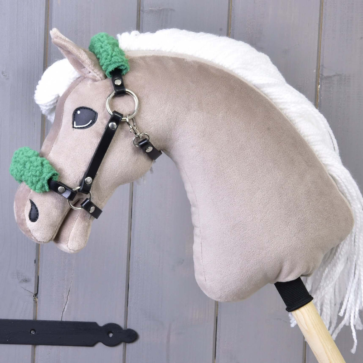 Bež Hobby Horse konj z belo grivo iz preje nosi črno oglavko iz umetnega usnja z zelenim krznom na nosniku in zatilnem jermenu