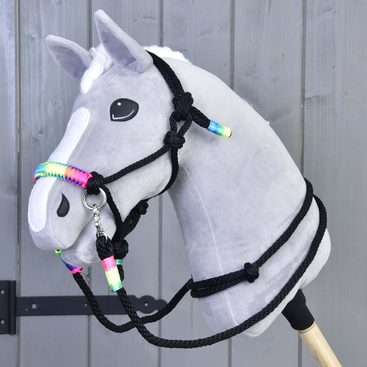 Siv Hobby Horse konj z belo liso, s črno vrvično oglavko in vajetmi z mavričnimi ovoji, ob sivi leseni steni