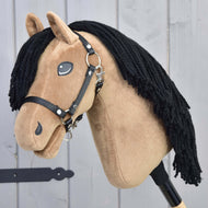 LarDen Hobby Horse Samantha s črno oglavko, plišasto glavo peščene barve in dolgo črno grivo iz preje, ob sivi leseni steni