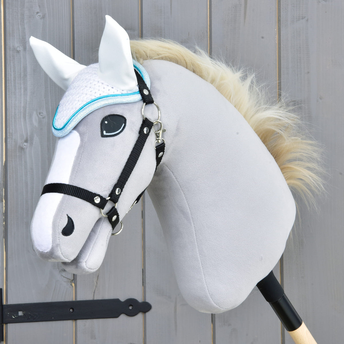 Svetlo siv Hobby Horse konj s črno oglavko in smetanasto grivo nosi bele kvačkane naušnike s turkizno obrobo