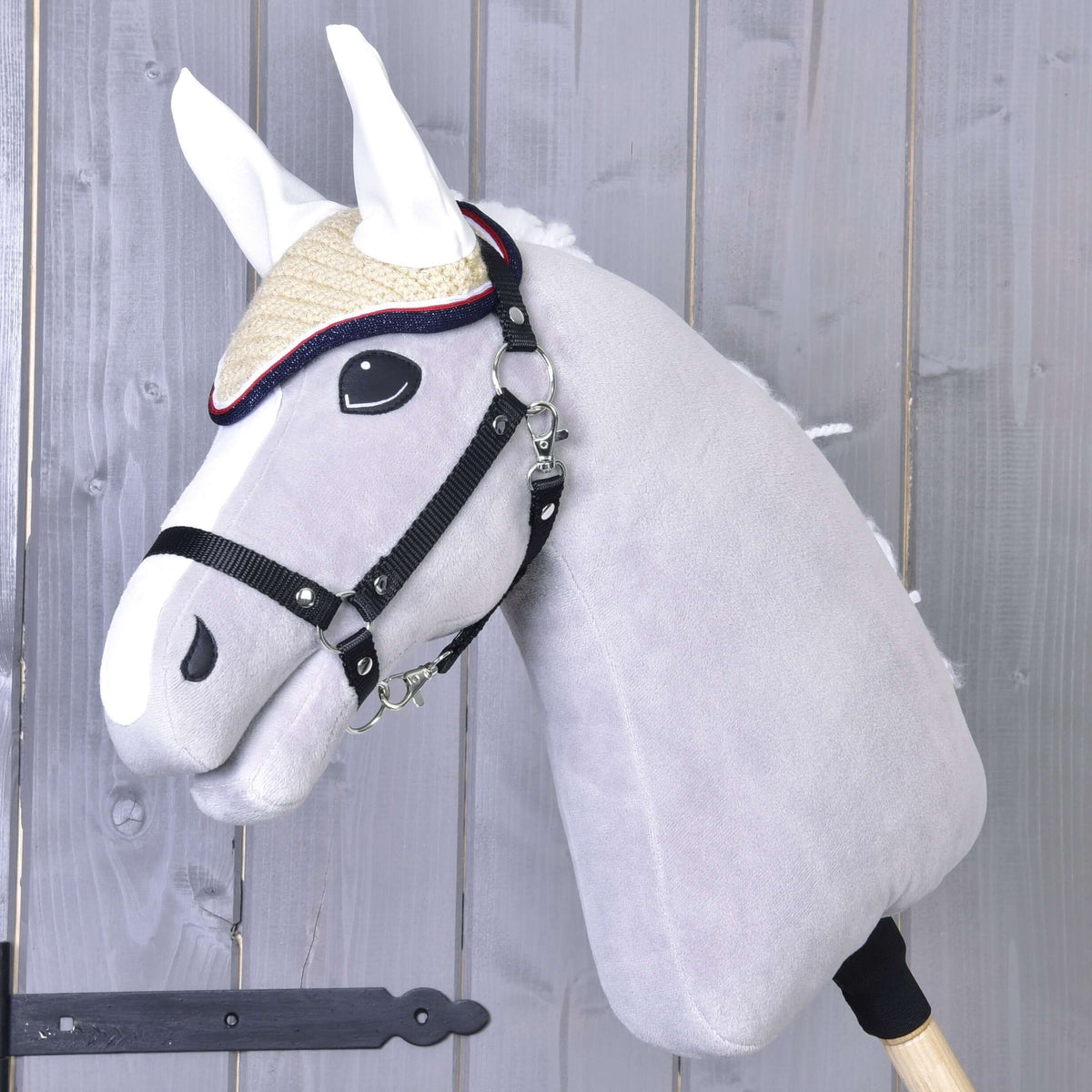 Siv Hobby Horse konj s črno oglavko nosi smetanaste kvačkane naušnike Nicol z belimi ušesi in rdeče-temno modrim robom