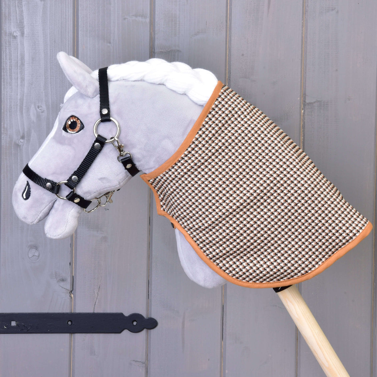 Siv plišast Hobby Horse konj s črno oglavko nosi pregrinjalo, obrnjeno na rjavo stran z vzorcem pepita, ob sivi steni