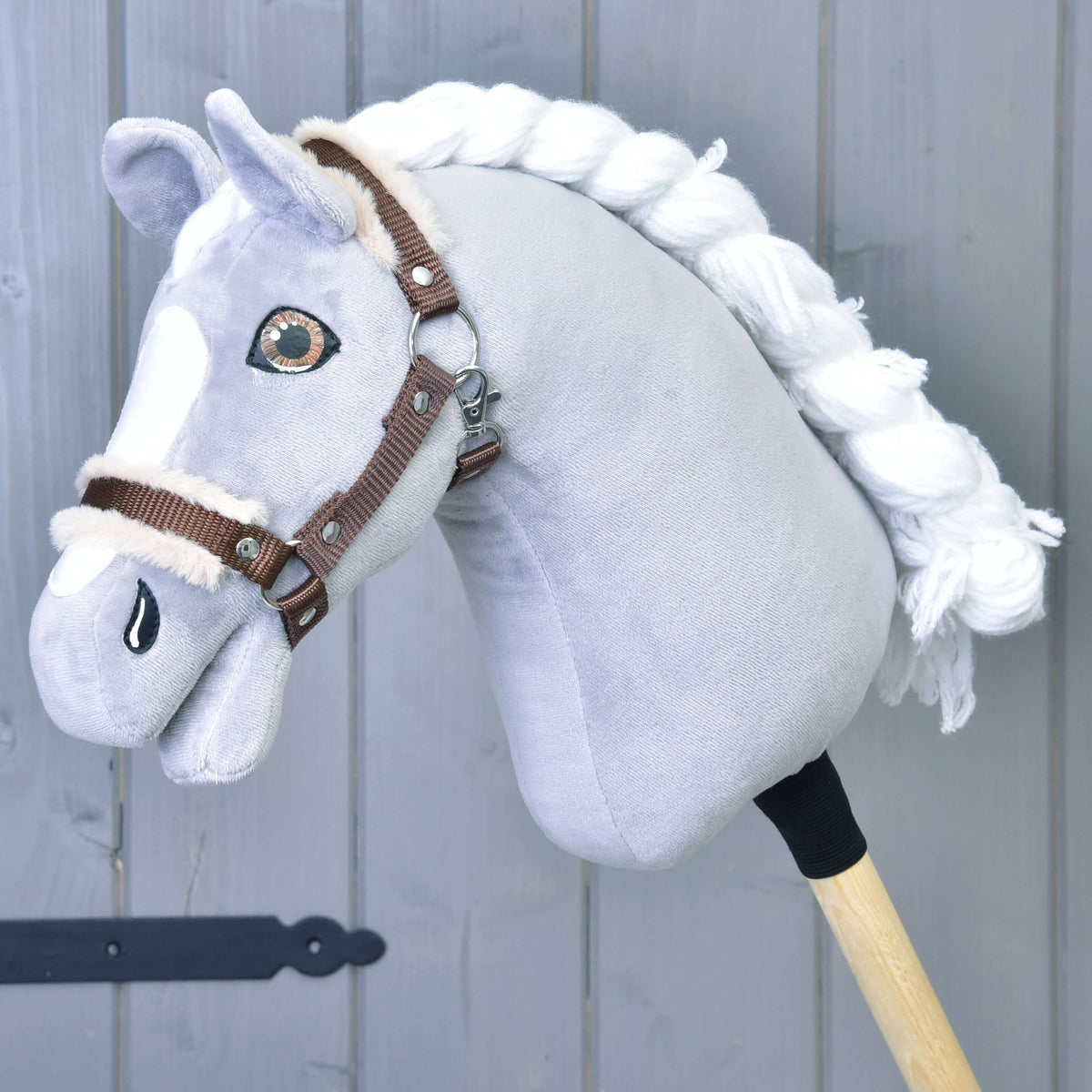 Siv plišast Hobby Horse konj s spleteno belo grivo in rjavimi očmi nosi rjavo oglavko z bež krznom