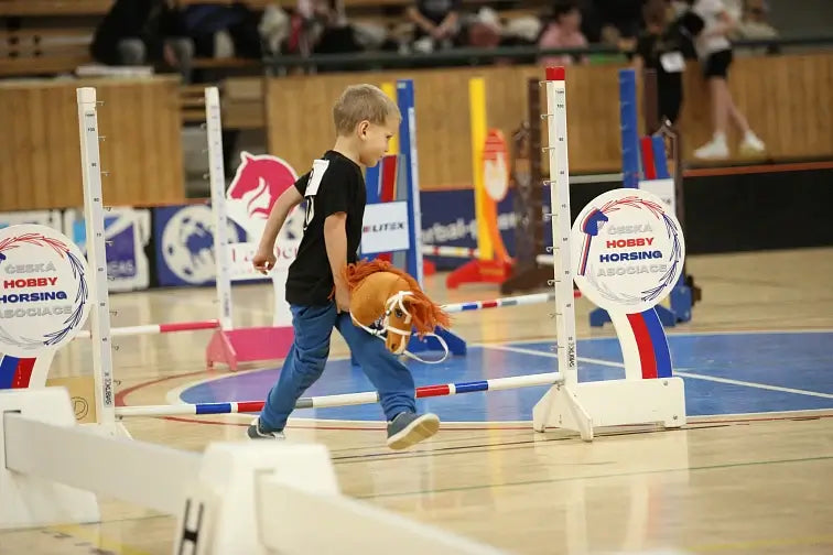 Deček sodeluje na Hobby Horse tekmovanju in hodi s konjem igračo po tleh telovadnice, obdan s pisanimi ovirami za skakanje in napisi tekmovanja v športu Hobby Horsing.