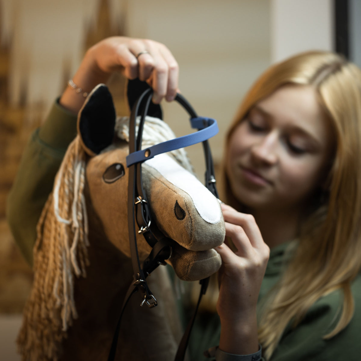 Svetlolasa deklica namešča temno rjavo uzdo z modrim čelnikom na svetlo rjavega Hobby Horse konja s svetlo grivo iz preje