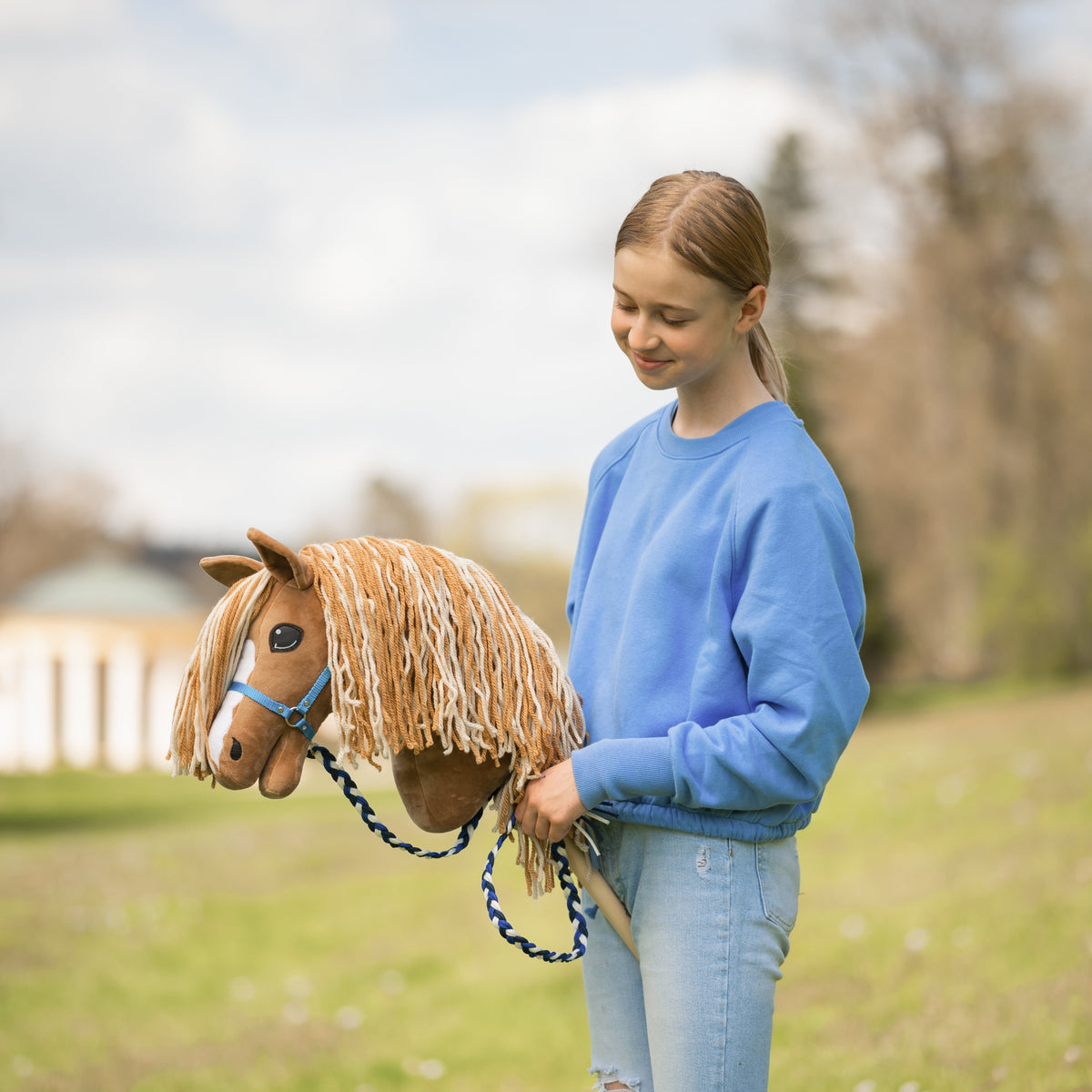 Deklica v modrem puloverju se nasmiha Hobby Horse konju, lisjaku z dolgo svetlo grivo, modro oglavko in pletenim povodcem