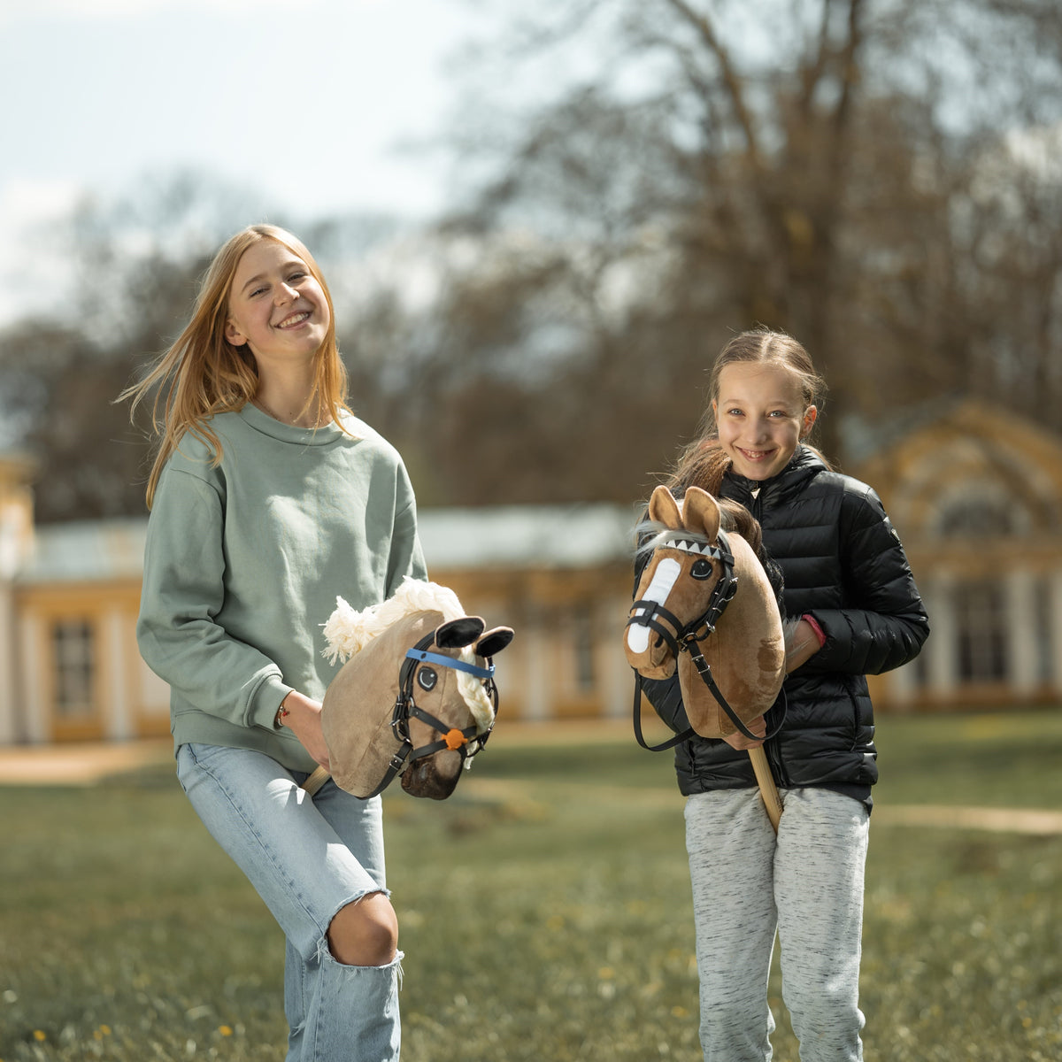 Dve nasmejani deklici v parku z bež in svetlo rjavim Hobby Horse konjem v črnih uzdah, zadaj rumena stavba