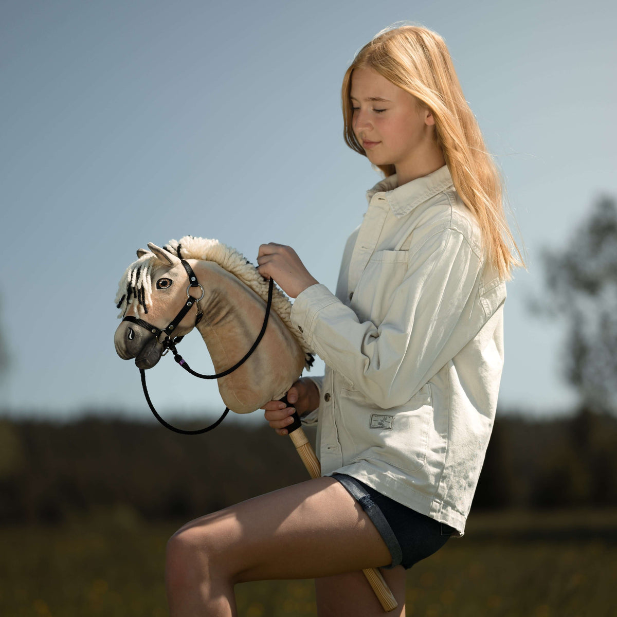 Svetlolasa deklica v kremni jeans jakni drži Hobby Horse konja, plavkastega rjavca s črno uzdo in vajetmi, pred modrim nebom