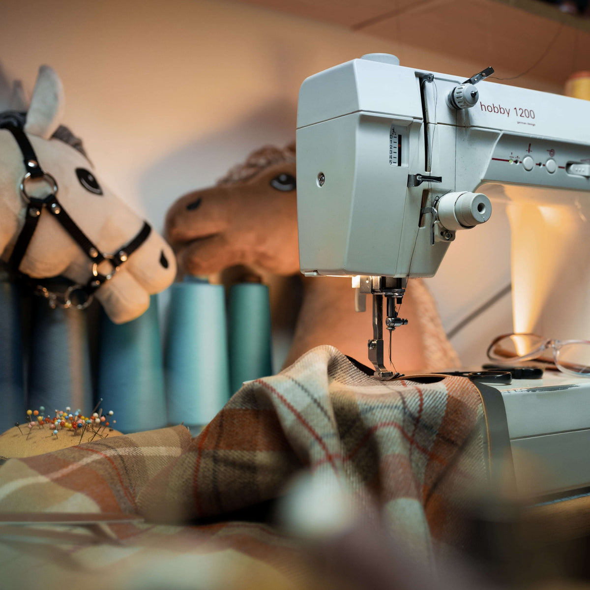 Handmade quality: a sewing machine stitches checked fabric, with teal thread and plush Hobby Horse heads behind