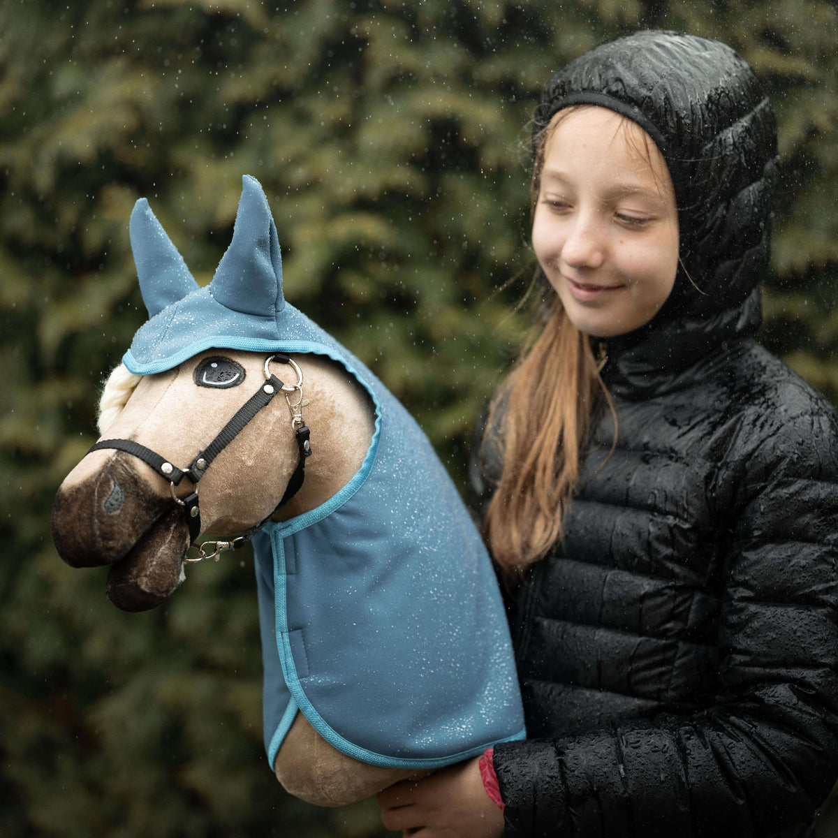 Smiling girl in a black puffer jacket in the rain with a beige Hobby Horse wearing a blue glittery raincoat with ears