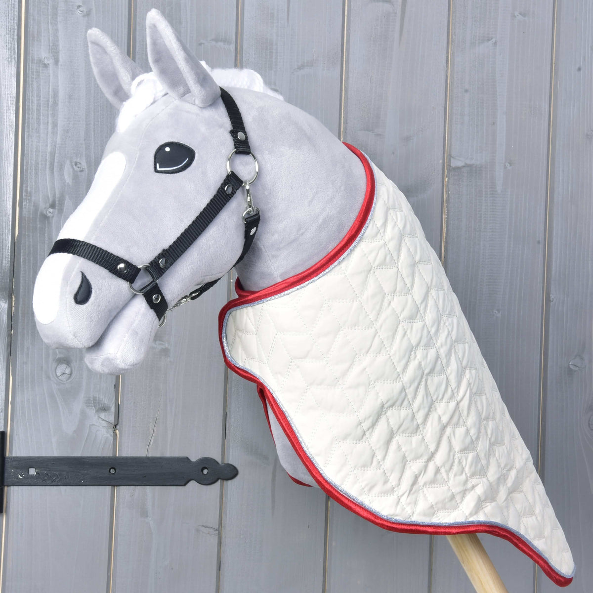 Siv Hobby Horse konj s črno oglavko, oblečen v kremno prešito transportno pregrinjalo z rdečo obrobo čez vrat