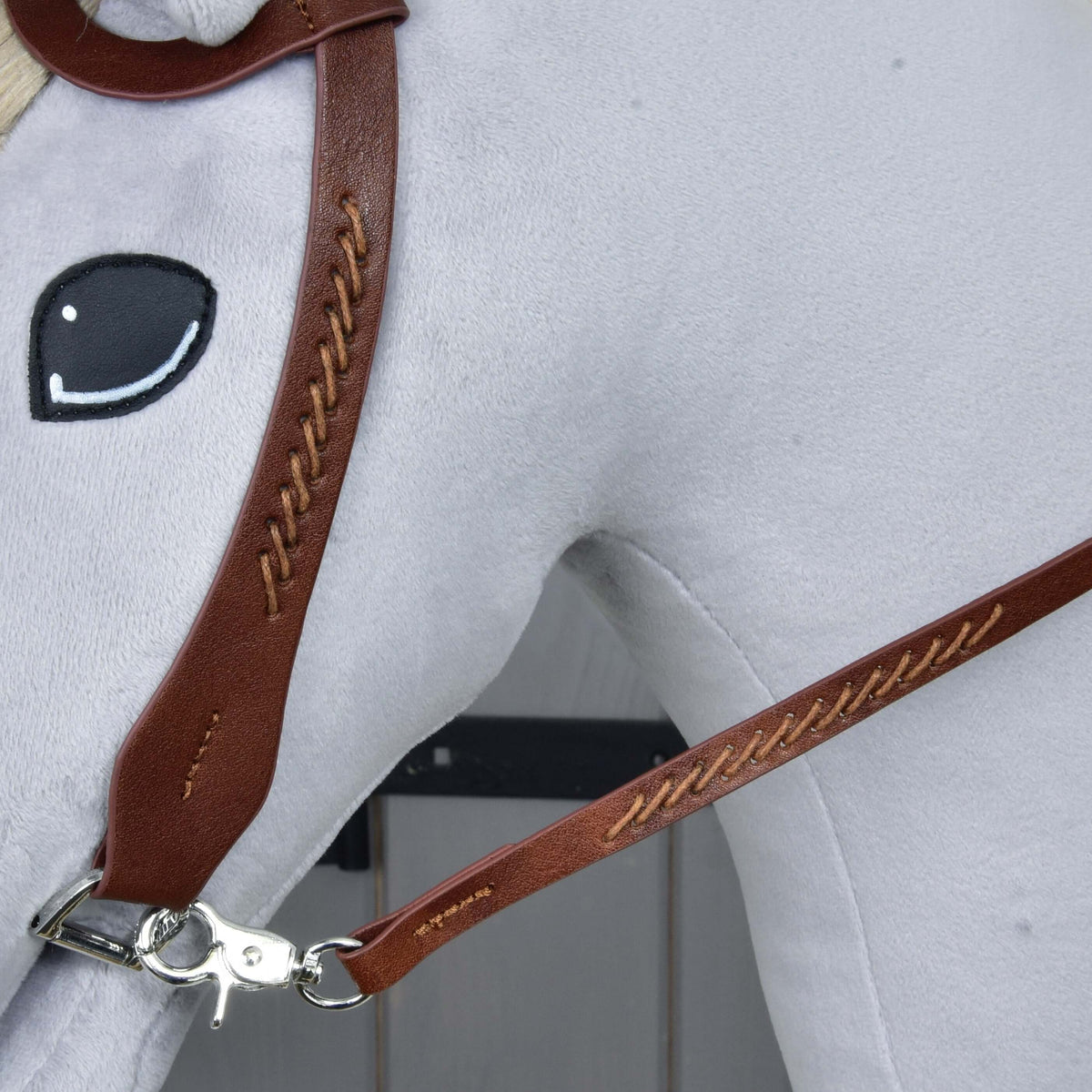 Close-up of the cheekpiece and rein with matching tan cross-stitching, joined by a silver snap hook on the grey plush head