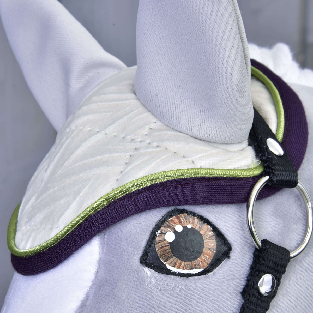 Detail of the lime green piping and aubergine binding on the ivory ear bonnet, just above the amber eye of a grey Hobby Horse