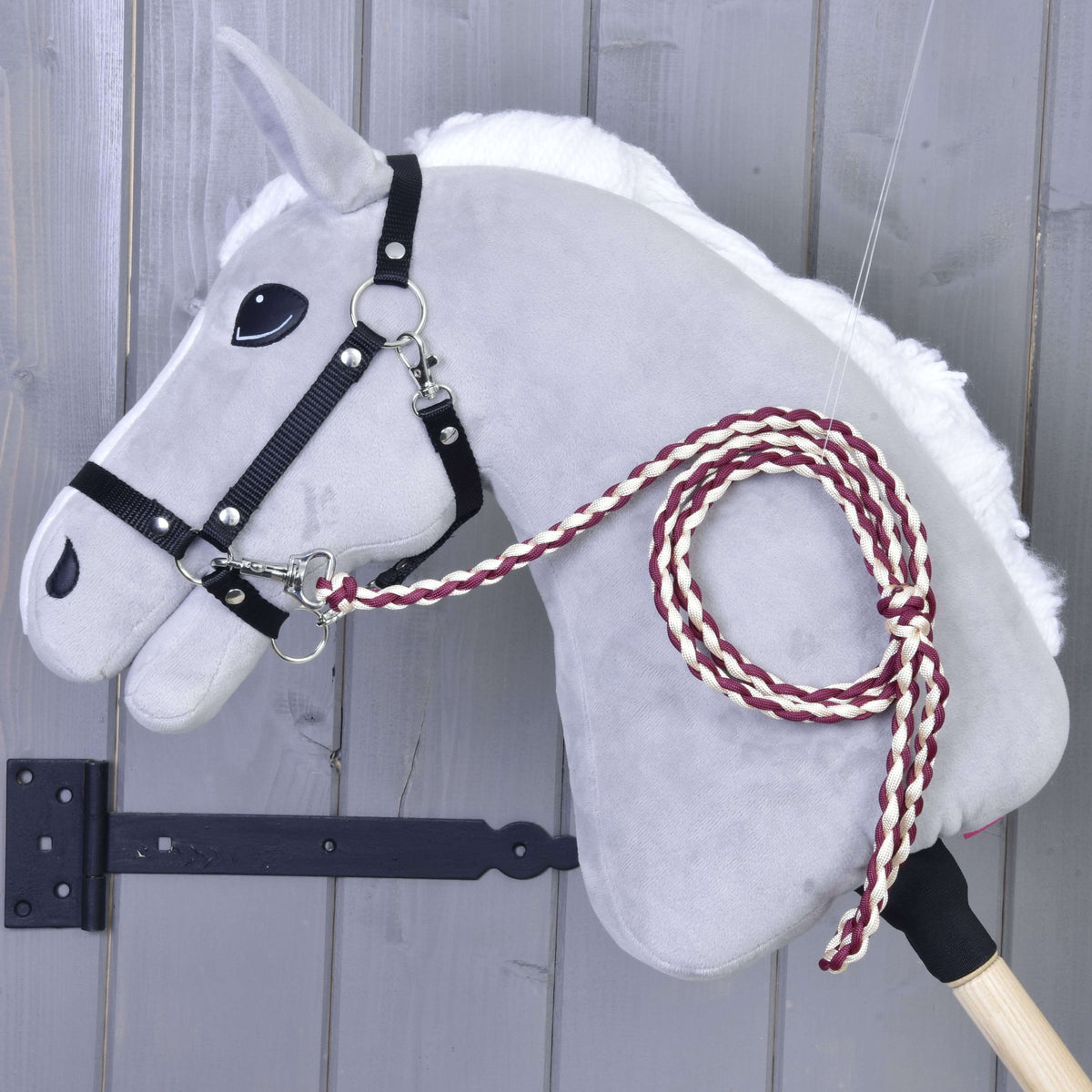 Pleten povodec v bordo in kremni barvi, pripet na črno oglavko sivega Hobby Horse konja ob sivi leseni steni