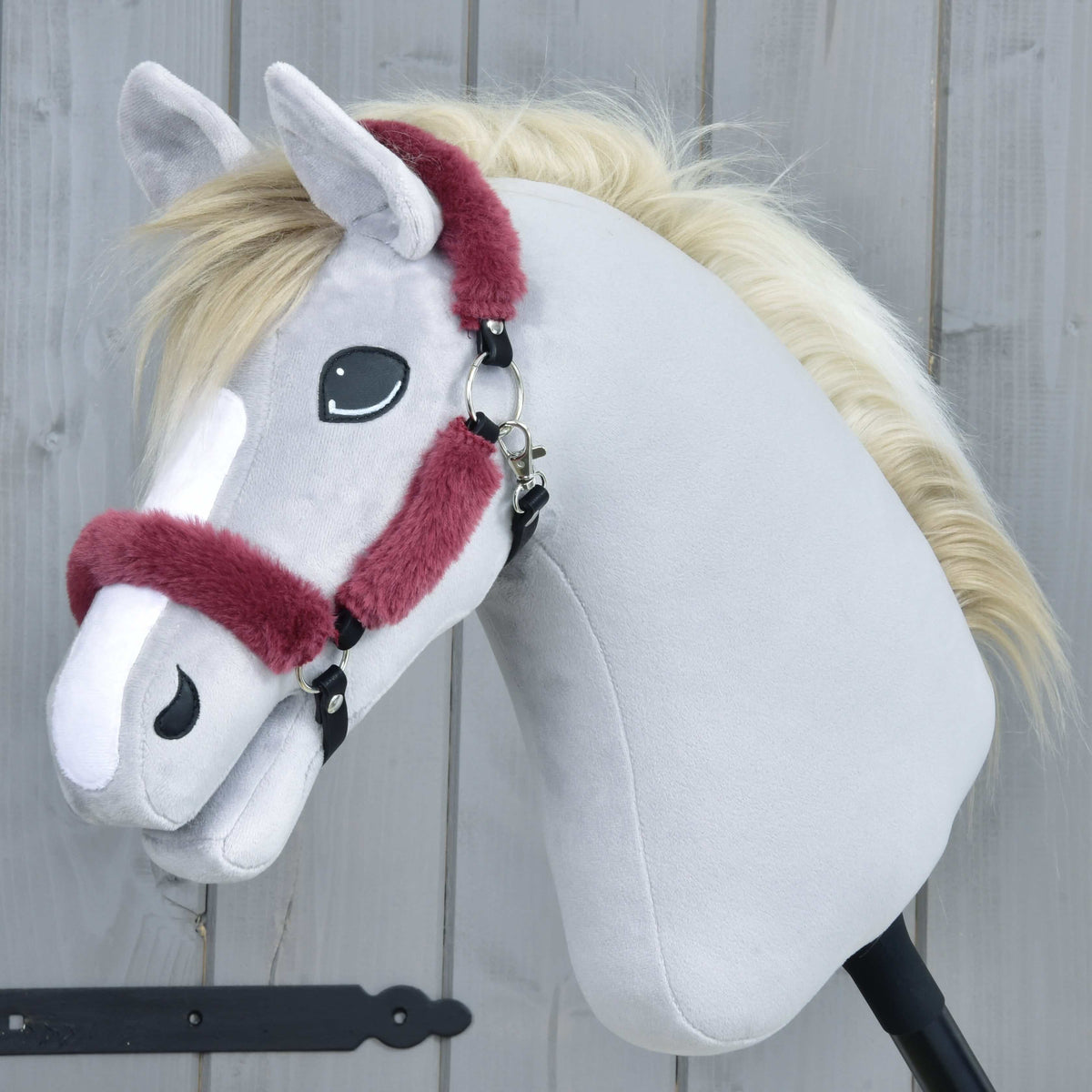 Grey plush Hobby Horse with a cream mane wearing the maroon fur halter, side view against a grey wooden wall