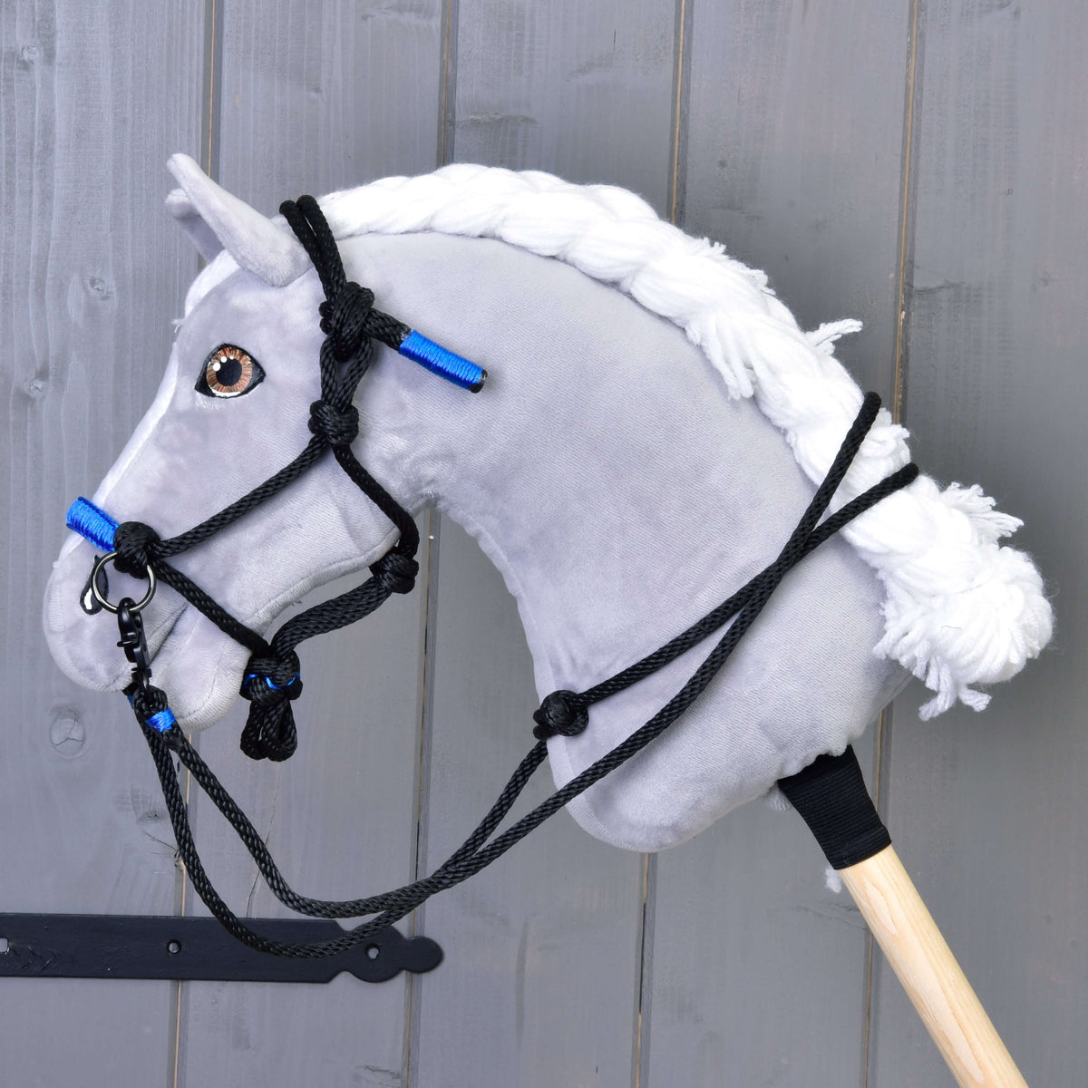Siv plišast Hobby Horse konj s spleteno belo grivo nosi črno-modro vrvično oglavko s črnimi vrvičnimi vajetmi