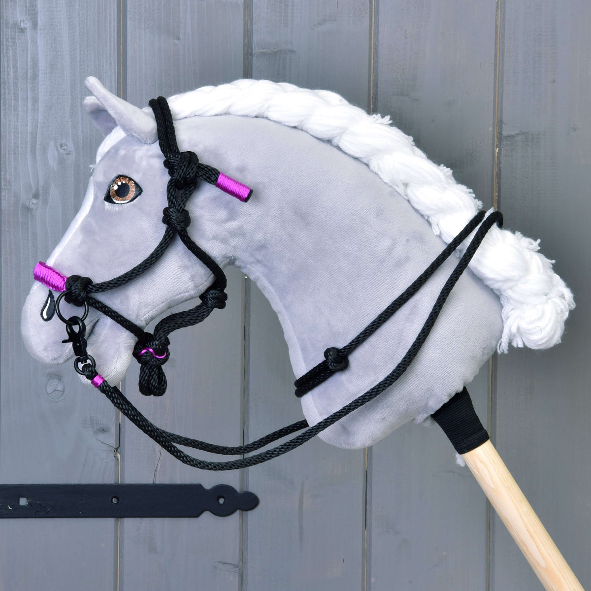 Siv plišast Hobby Horse konj s spleteno belo grivo nosi črno-vijolično vrvično oglavko s črnimi vrvičnimi vajetmi