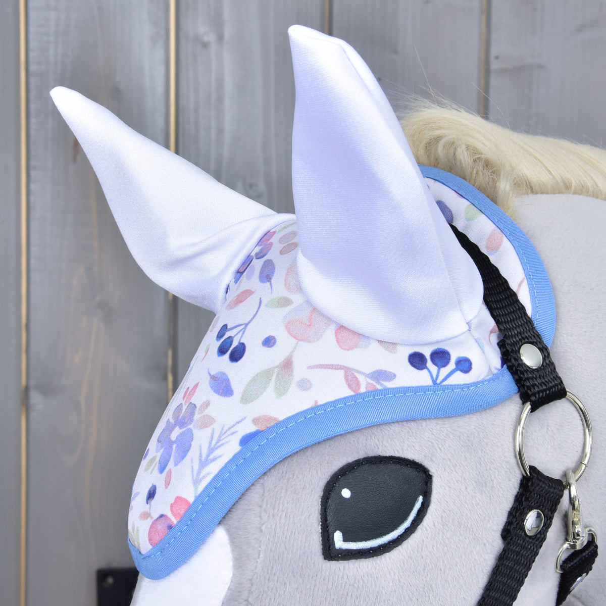 Close-up of the floral ear bonnet with shiny white ears and blue trim on a grey Hobby Horse beside the black halter