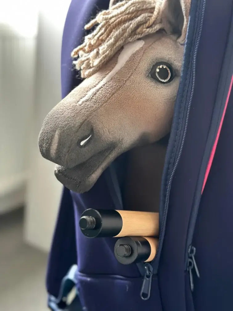 Light brown Hobby Horse with a beige yarn mane peeking out of a navy backpack beside a two-part wooden stick
